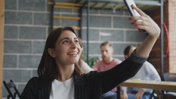 Mujer emprendedora alegre sosteniendo teléfono inteligente y tener videollamada en línea mientras sus colegas trabajan en la oficina moderna — Vídeos de Stock