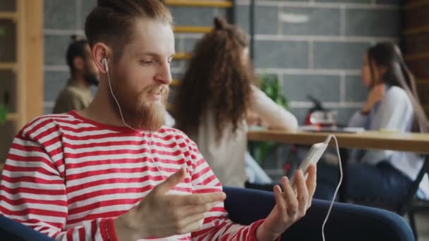 Hipster hombre tienen videollamada en línea utilizando el teléfono inteligente durante el descanso, mientras que sus colegas que trabajan en la oficina moderna — Vídeos de Stock