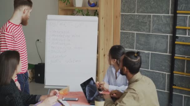 Joven hipster ceo masculino explicando plan de negocio de puesta en marcha en el rotafolio a los empleados en la oficina loft moderna — Vídeos de Stock