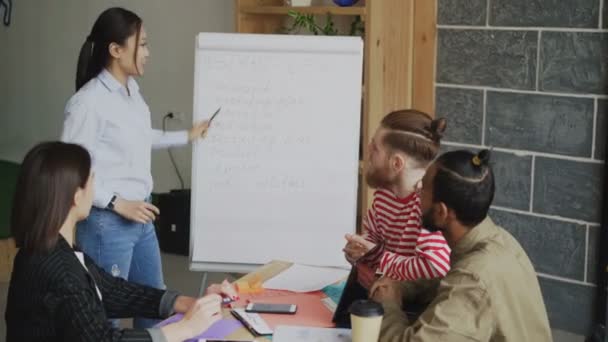 Gai asiatique fille expliquer start-up business plan sur tableau à feuilles mobiles pour collègues dans moderne loft bureau — Video