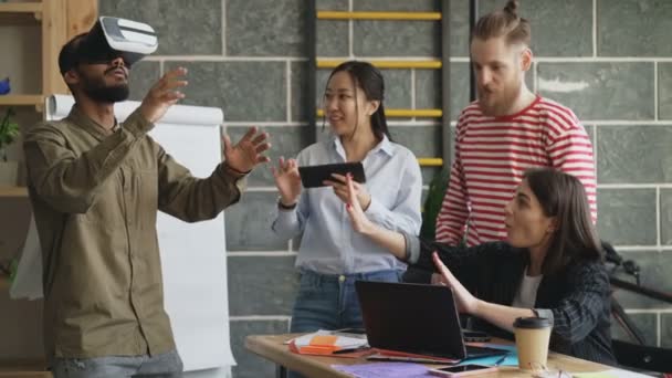 Mult ethnic team testing VR game simulator at modern office. African american man trying play virtual reality headset while asian girl using digital tablet — Stock Video