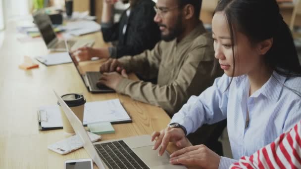 Több-etnikumú csapat asztalnál indítási projekten dolgozó munkatársak. Ázsiai koncentrált nő gépelés-ra laptop számítógép modern loft hivatalban — Stock videók