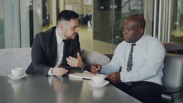 Dos socios multiétnicos en ropa formal mirando la tableta digital y discutiendo sus ideas de negocio en una mesa con bloc de notas y una taza de café en un café de moda — Vídeos de Stock