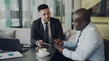 Sözleşmeleri seyir ve onların masada dizüstü bilgisayar ve havadar kafede grafikler ile şarkı iki iddialı iş ortağı. Afrika kökenli Amerikalı işadamı ve beyaz adam el sallamak