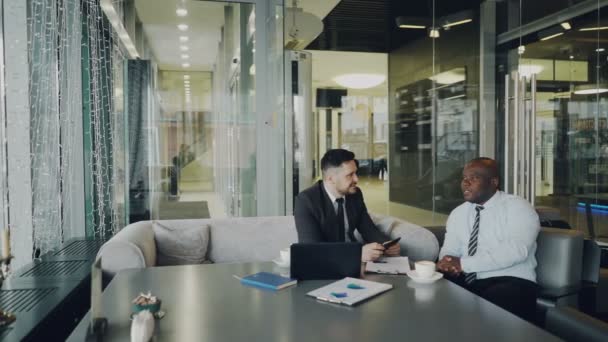 Dos emprendedores masculinos optimistas en ropa formal discutiendo su proyecto de startup en la cafetería. Empresarios afroamericanos y caucásicos conversando y sentados a la mesa — Vídeos de Stock