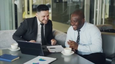 Dizüstü bilgisayar seyir ve camsı Cafe gülüyor iki çok etnik işadamları. Afrika kökenli Amerikalı işadamı ve eğleniyor ve şaka beyaz iş arkadaşları