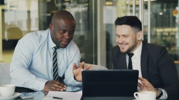 Dos colegas de negocios mirando el ordenador portátil y discutiendo sus proyectos en la oficina moderna con paredes de vidrio. Empresario barbudo y su compañero sentados a la mesa y hablando — Vídeo de stock