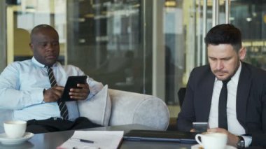 İki iş arkadaşları resmi içinde oturan ve onların smartphone ve tablet toplantı sırasında camsı kafede kullanarak giymek. Internet bağımlılığı, teknoloji ve mobil ofis kavramı