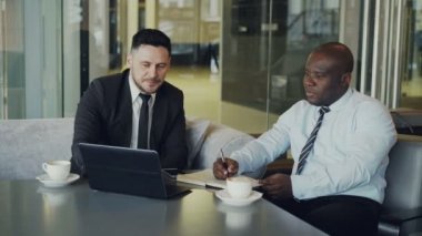 İki neşeli işadamları proje modern café'tartışıyor. Afrika kökenli Amerikalı girişimci dizüstü bilgisayar ve dizüstü bilgisayar işaret iş elbiseli beyaz ortağı yazma