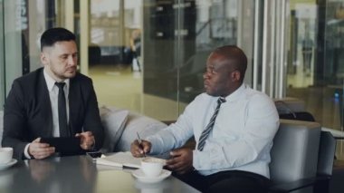 Pan hırslı girişimcilerin başlangıç projesi modern cam gibi lobisinde tartışıyor. Afrika kökenli Amerikalı işadamı defter ve beyaz ortağı iş elbiseli dijital tablet kullanarak yazma