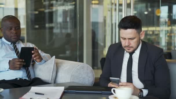 Dois empresários multi-étnicos em roupas formais sentados e usando seu smartphone e tablet em café vítreo durante a hora do almoço — Vídeo de Stock