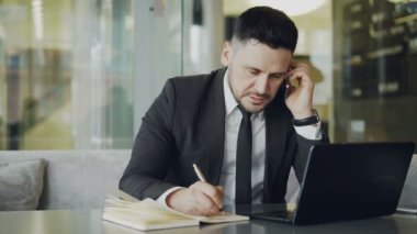 Oturup, onun smartphone üzerinde konuşmak ve iş öğle yemeği zamanı sırasında camsı kafede dizüstü bakarak süre onun not defteri bilgileri yazma konsantre beyaz iş adamı