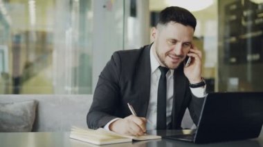 Oturan, gülümseyen, smartphone ortağıyla konuşmak ve kafede dizüstü bakarak süre not defteri bilgileri yazma mutlu beyaz işadamı