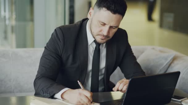 Συμπυκνωμένο γενειοφόρος Καυκάσιος επιχειρηματίας, κάθονται και το γράψιμο κάτω επιχειρηματικές πληροφορίες στο σημειωματάριο του ενώ κοιτάζοντας το laptop στο mod καφέ — Αρχείο Βίντεο