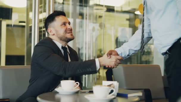 Glad kaukasiska affärsman i formella kläder leende och skakar hand med sin afrikanska amerikanska partner att ta plats i mod café under lunchen. — Stockvideo