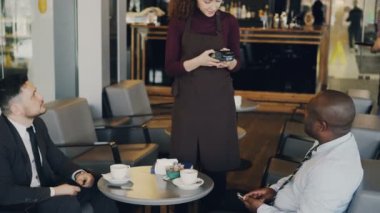 İki çok ırklı iş adamları formal giyim modern kafede smartphone kullanarak online fatura ödeme. Ayakta ve taşınabilir nakit terminal tutmak genç garson.