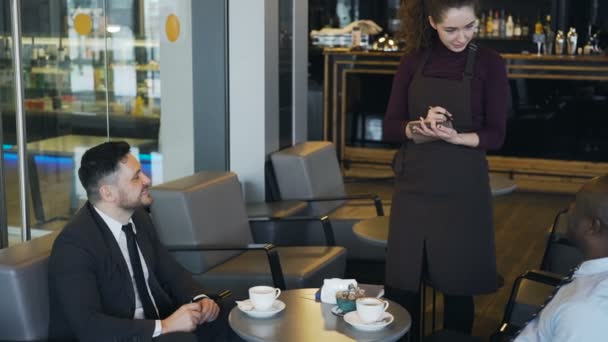 Zwei multiethnische Geschäftsleute in formeller Kleidung sitzen in der Mittagspause lächelnd in einem stilvollen Café und sorgen für Ordnung. junge Kellnerin schreibt ihr Geschirr auf — Stockvideo