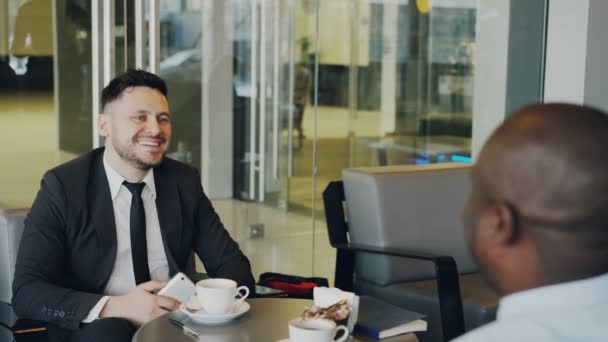 Homem de negócios caucasiano barbudo sorridente sentado com seu parceiro afro-americano e pagando conta on-line usando seu smartphone no café. Garçonete alegre usando terminal de caixa . — Vídeo de Stock