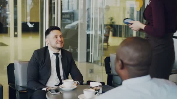 Felice uomo d'affari caucasico barbuto seduto con il suo collega afroamericano e pagare la bolletta online utilizzando il suo smartphone in caffè. Cameriera sorridente utilizzando il terminale di cassa . — Video Stock