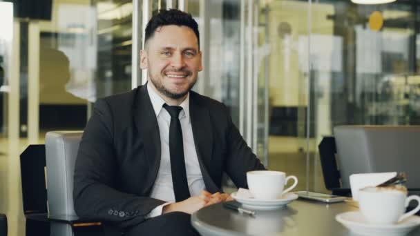 Retrato de feliz hombre de negocios caucásico barbudo en ropa formal sentado, bebiendo café y sonriendo en la cafetería de cristal mientras almorzaba. Smartphone está en su mesa — Vídeos de Stock