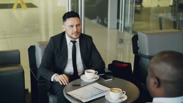 Heureux homme d'affaires caucasien barbu souriant, gesticulant, buvant du café et discutant du rapport financier de la start-up avec son partenaire afro-américain dans le café — Video