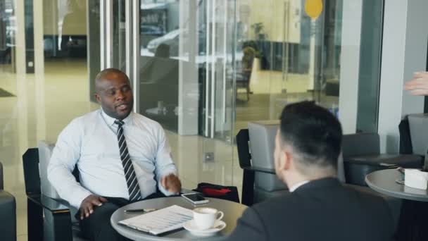 Alegre hombre de negocios afroamericano con ropa formal sonriendo y discutiendo el informe financiero con su socio caucásico en la cafetería. Camarera joven trayendo una taza de café . — Vídeos de Stock