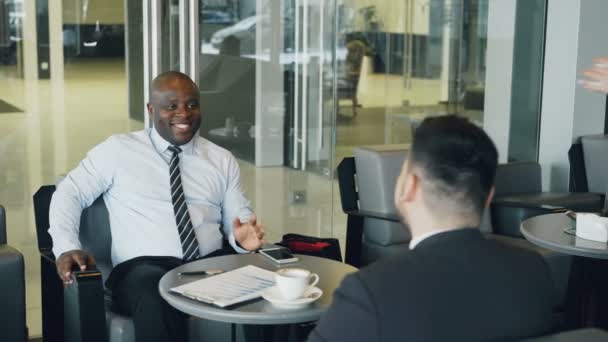 Empresário afro-americano bem sucedido em roupas formais sorrindo e discutindo relatório financeiro com seu colega caucasiano no café. Garçonete jovem trazendo xícara de café — Vídeo de Stock