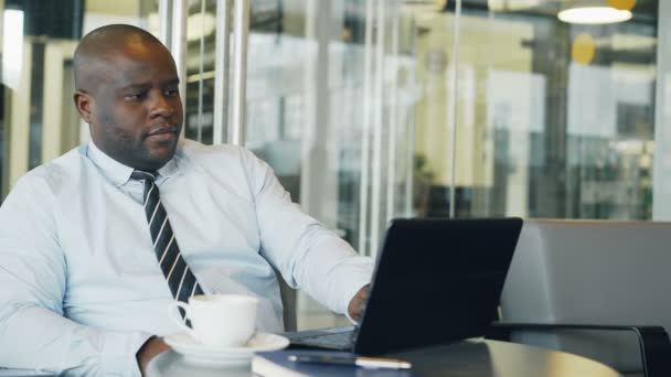 Hombre de negocios afroamericano pensando, imprimiendo y trabajando en su computadora portátil en una cafetería acristalada durante la pausa para el almuerzo. Está haciendo una lluvia de ideas innovadoras. . — Vídeos de Stock