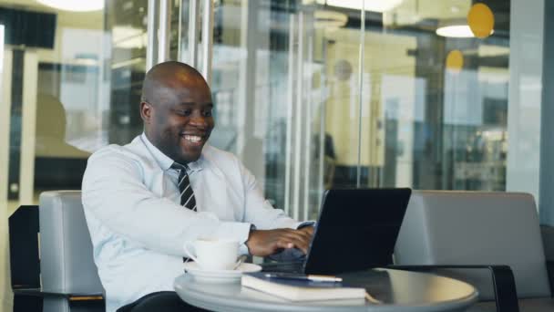 Успішний бізнесмен афроамериканець, використовуючи ноутбук комп'ютер, який отримує хороші повідомлення і стати дуже схвильований, танцювальні, сидячи в сучасних кафе — стокове відео