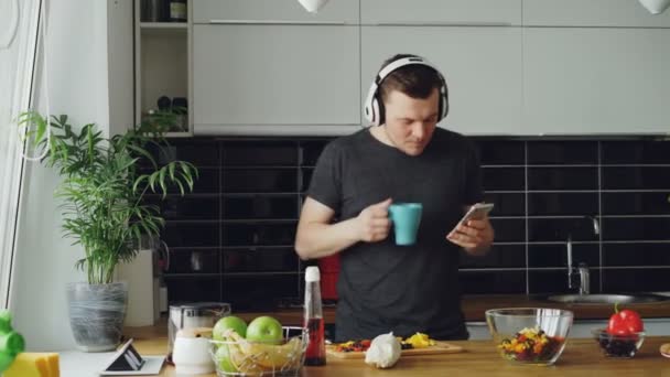 Veselý šťastný muž tančí a zpívá v kuchyni při surfování sociální média na jeho smartphone doma ráno — Stock video