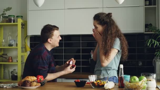 Молодой человек делает предложение своей девушке, пока она готовит дома на кухне. Он надел кольцо ей на палец и поцеловал ее. — стоковое видео