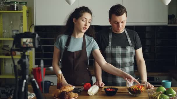 Casal atraente sorrindo gravando vídeo food vlog sobre cozinhar salada de feno na câmera digital na cozinha em casa. Vlogging, educação e conceito de mídia social — Vídeo de Stock