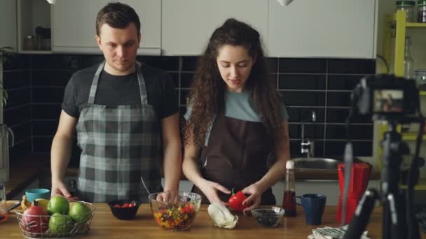 Весёлая привлекательная пара записывает видеоблог о здоровой готовке на цифровую камеру на кухне дома. Ведение блогов, образование и концепция социальных сетей — стоковое видео