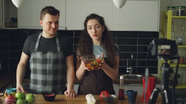 Casal atraente alegre gravando vídeo food vlog sobre culinária saudável na câmera digital na cozinha em casa. Vlogging e conceito de mídia social — Vídeo de Stock