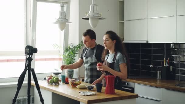 Lächelndes attraktives Paar, das Video über das Kochen von Salat mit der Digitalkamera in der heimischen Küche aufzeichnet. vlogging, Bildung und Social-Media-Konzept — Stockvideo