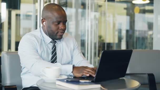 Empresário afro-americano em uso formal trabalhando e navegando nas mídias sociais no laptop enquanto ouve música com fones de ouvido em seus ouvidos em um café vítreo — Vídeo de Stock