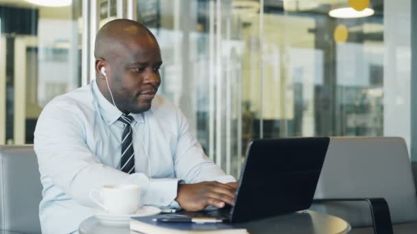 Afro-Amerikaanse ondernemer in formele kleding bezig met laptop terwijl u luistert naar muziek met oordopjes in zijn oren in een moderne café — Stockvideo