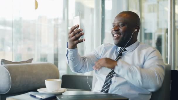 Hombre de negocios afroamericano en ropa formal riendo y hablando con su familia a través de chat de vídeo smartphone en la cafetería aireada. Él agita su mano alegremente . — Vídeos de Stock