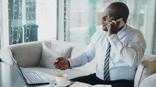 African American businessman talking on smartphone and putting down business information in his notepad sitting in modern cafe with laptop and coffee on his table. — Stock Video