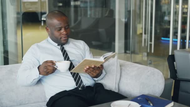 Slimme Afro-Amerikaanse zakenman in formele kleding lezen boek terwijl het drinken van koffie in glazig café tijdens lunchpauze — Stockvideo