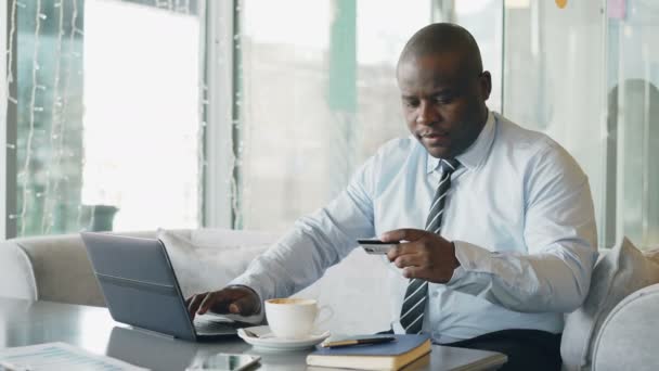 Sorridente uomo d'affari afro-americano in abiti formali pagare bolletta online utilizzando la carta di credito e laptop in caffè alla moda durante il pranzo respirare . — Video Stock