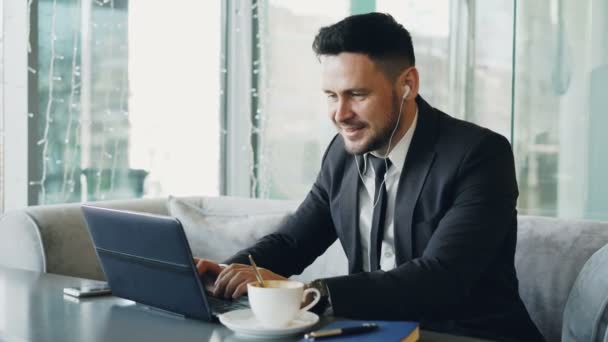 Empreendedor caucasiano feliz em roupas formais digitando em seu laptop enquanto ouve música com fones de ouvido em seus ouvidos em um café moderno durante o almoço — Vídeo de Stock
