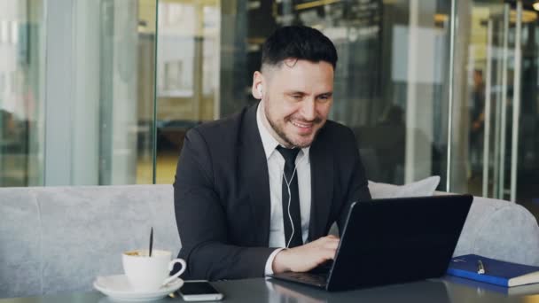 Feliz empresario caucásico en ropa formal escribiendo en su computadora portátil mientras escucha música con auriculares en sus oídos en un café moderno durante el almuerzo — Vídeos de Stock