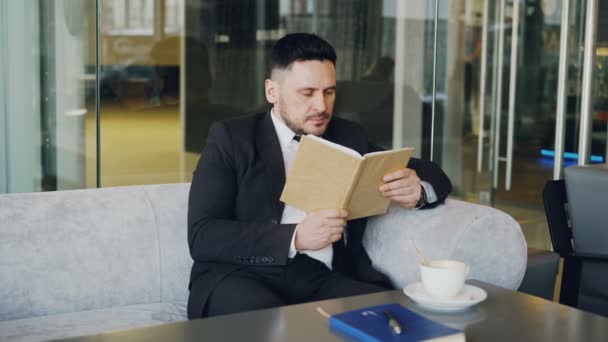 Inteligente hombre de negocios caucásico en ropa formal lectura libro de texto con y beber café en elegante cafetería en la pausa para el almuerzo — Vídeo de stock