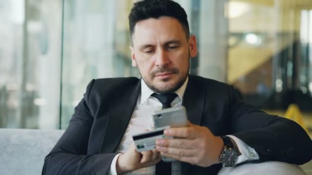 Uomo d'affari caucasico sorridente in giacca e cravatta con carta di credito online banking holding e smartphone in mano in un moderno caffè durante la pausa pranzo — Video Stock