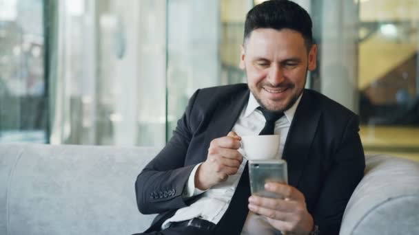 Glücklicher kaukasischer Geschäftsmann in formeller Kleidung, der in der Mittagspause im luftigen Café Kaffee trinkt und auf seinen Smartphone-Bildschirm lächelt — Stockvideo