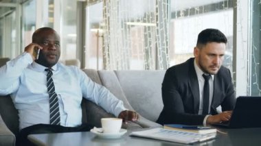 Beyaz işadamı resmi elbise diz üstü ve Afrika kökenli Amerikalı meslektaşı smartphone kafede konuşurken iş bilgilerini yazdırma.