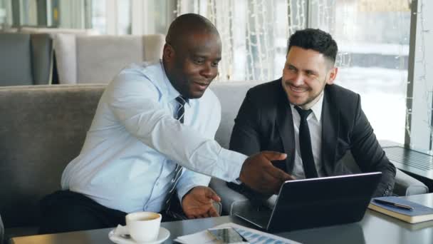 Vrolijke Afro-Amerikaanse zakenman in formele kleding bespreken zakelijk project met zijn Kaukasische collega op zijn laptop in café — Stockvideo