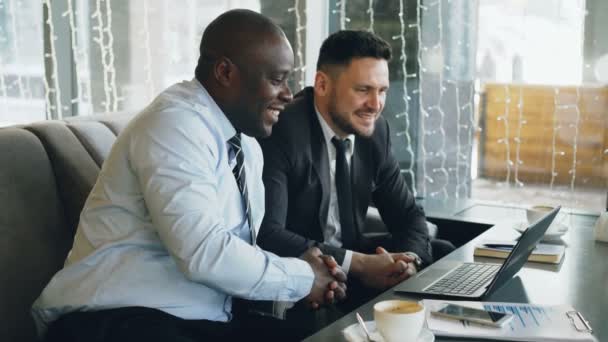 Fröhlich afrikanisch-amerikanischer Geschäftsmann in formeller Kleidung diskutiert Geschäftsprojekt mit seinem kaukasischen Kollegen auf seinem Laptop im Café — Stockvideo