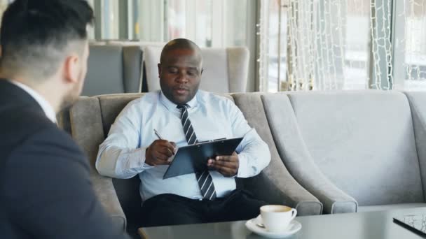 Responsabile delle risorse umane afroamericane che fa colloqui di lavoro con un giovane in giacca e cravatta e guarda la sua candidatura per il curriculum in un bar moderno durante una pausa caffè — Video Stock